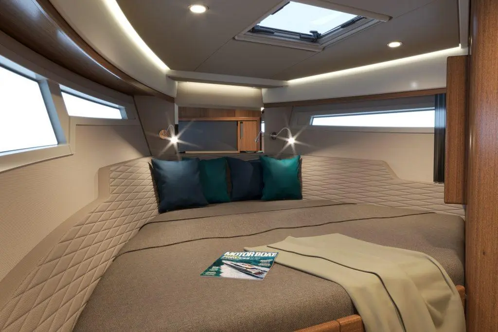 Modern boat cabin featuring a double bed with cushions, reading lights, two windows, and a magazine titled "Motor Boat.